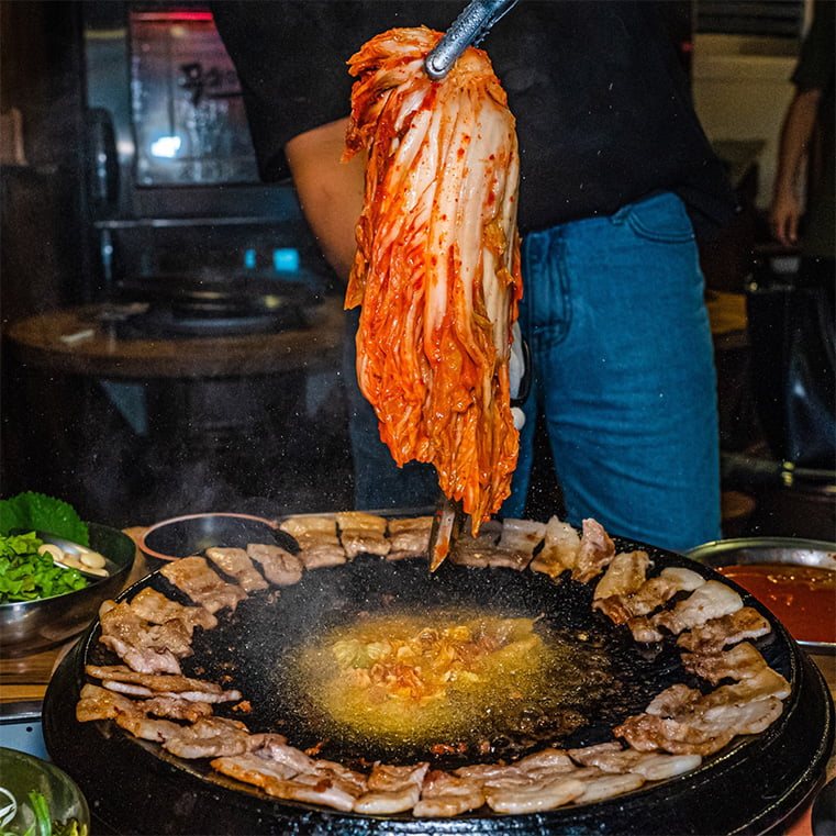 삼겹살을 둘러 구운 무쇠 뚜껑 불판 가운데, 고인 돼지기름에 통김치를 올리는 모습